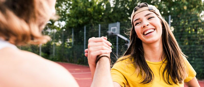 Young woman with a hat holding a basketball and shaking hand with a friend on outdoor basketball court while smiling | LISTERINE