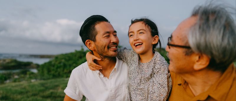 Young girl sitting on her father's lap next to her grandfather while smiling outdoors