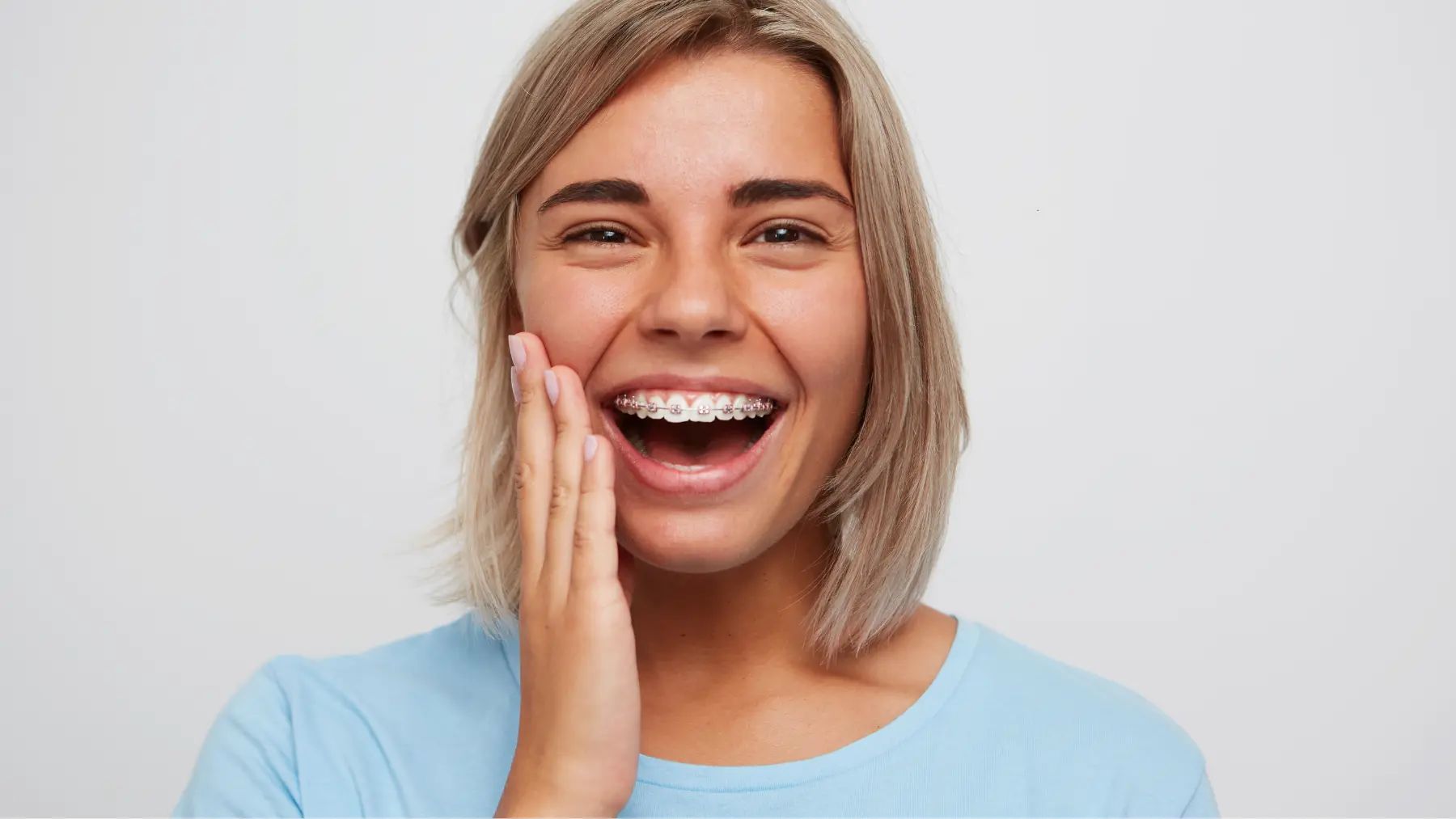 A woman with braces smiles radiantly while touching her cheek, showing a moment without tooth pain