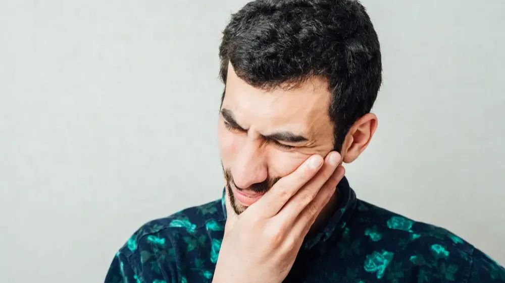 Un homme avec le visage crispé de douleur, la main sur la joue gauche.