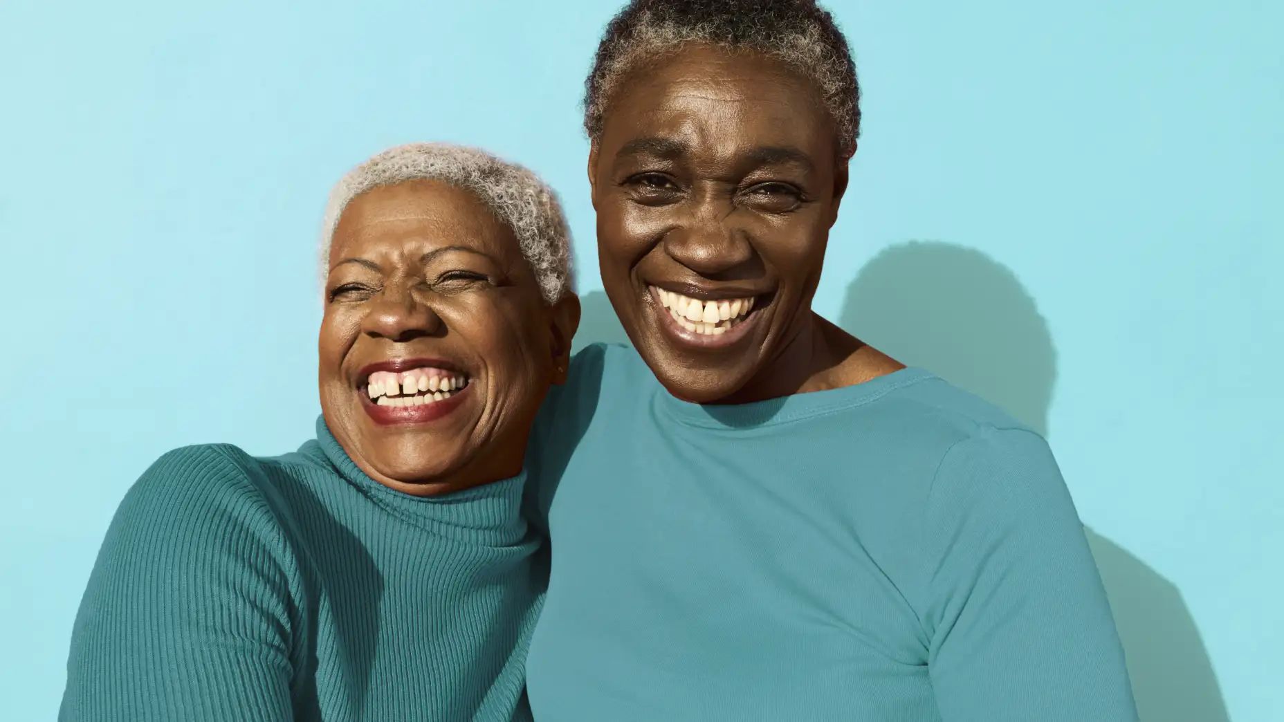 Two older women smiling together, standing against a vibrant blue background, swish for success
