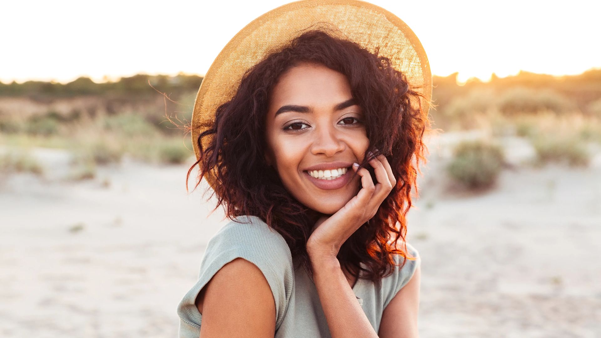 Une femme souriante sur la plage avec un chapeau sur la tête.