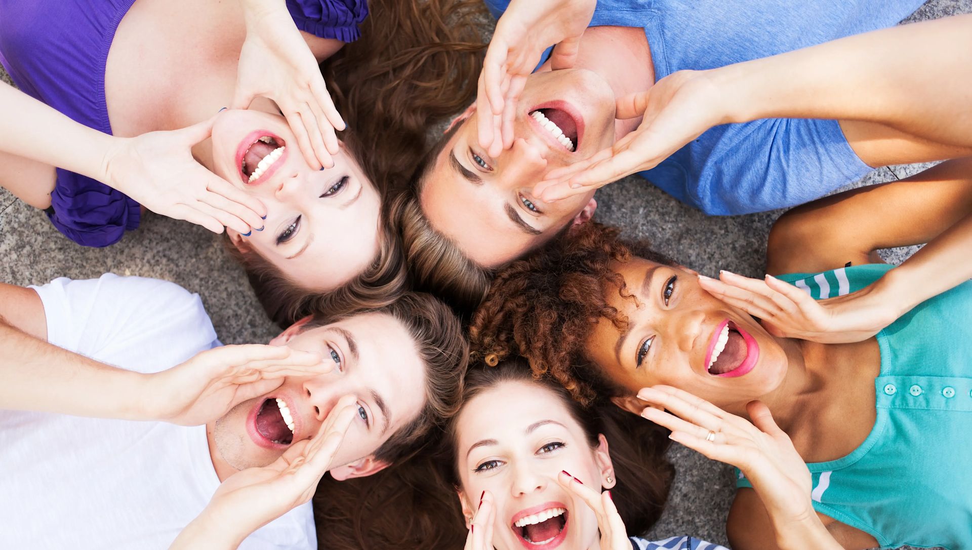 Eine Gruppe von heranwachsenden Jungen und Mädchen, die ihre Münder lächelnd geöffnet haben und auf dem Boden liegen. Die Hände sind so positioniert, als würden sie alle jemanden rufen.