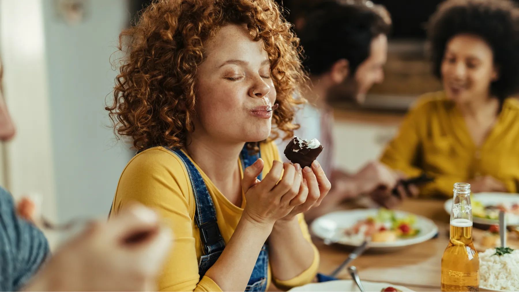 A woman eats cake at a table with friends, sharing smiles and laughter in a warm, social setting