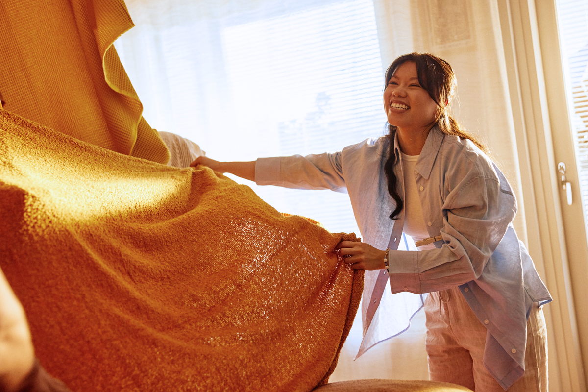 Nuori tummatukkainen nainen keltaisen huovan kanssa SATOn vuokra-asunnossa. Auringonvalo osuu huopaan, nainen nauraa iloisesti.