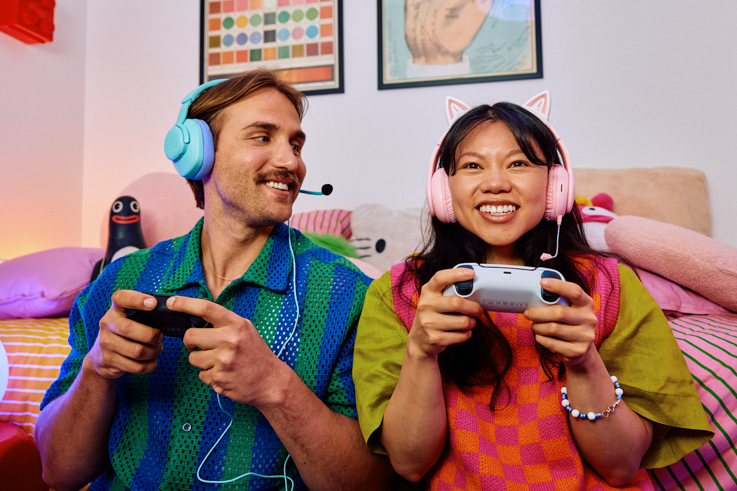 Kaksi iloista ihmistä pelaamassa konsolipeliä kuulokkeet korvillaan. Huoneen sisustus värikäs ja kotoisa. Taustalla kaksi taulua.