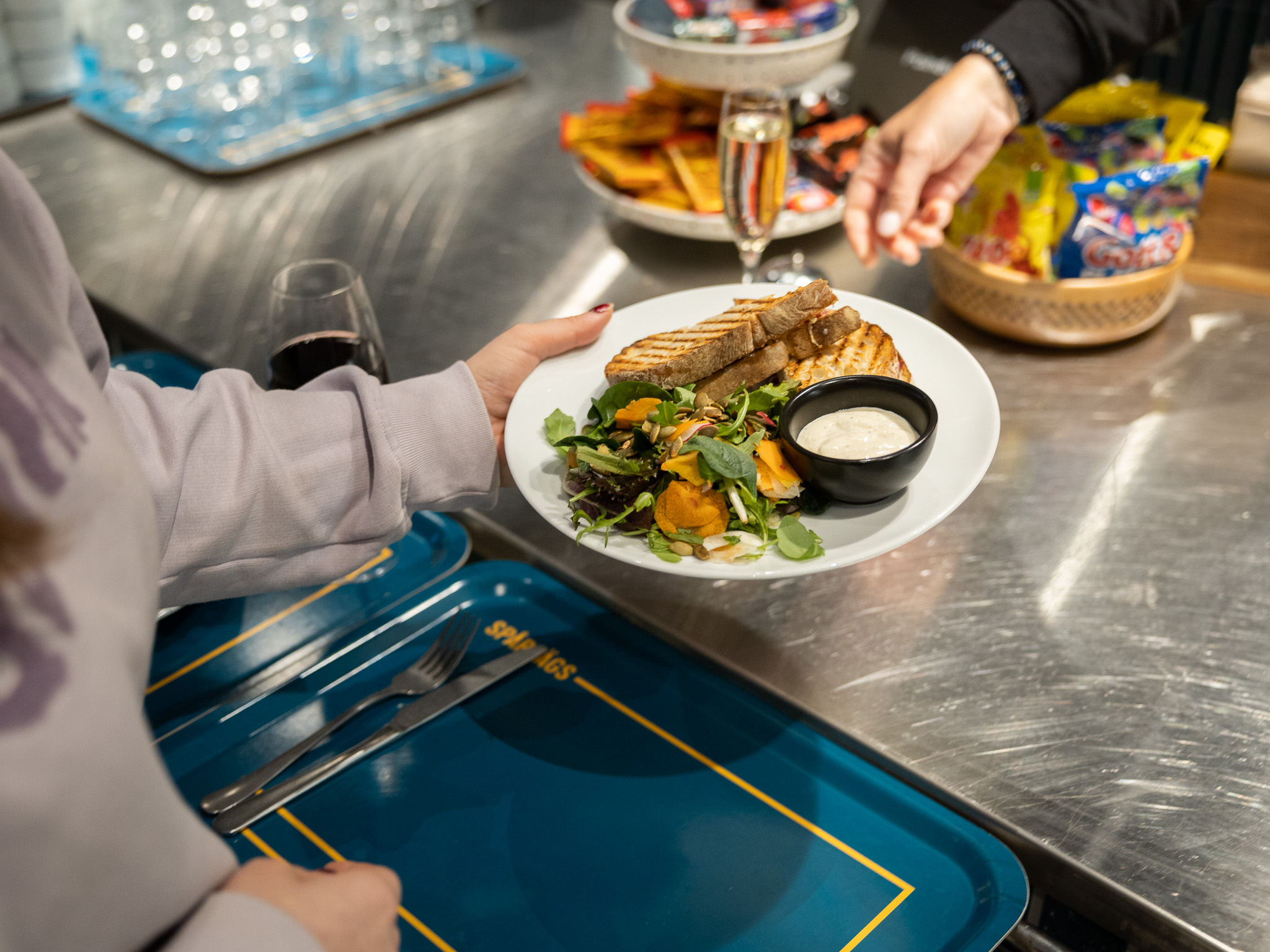 Två personer står vid en cafédisk. Personen i vänstra hörnet har en ljusrosa tröja på sig och håller i en tallrik med sallad, grillade mackor och sås på. Personen tar emot tallriken från en annan person som bär en svart tröja. Det är enbart personens högra hand som syns i bild. I förgrunden syns ett antal chokladkakor och godispåsar. Ett glas med bubbel står vid godiset. Nedanför personen som håller i en tallrik med mat ser vi två blåa brickor med gula detaljer på samt ett glas rött vin precis bakom personens arm.