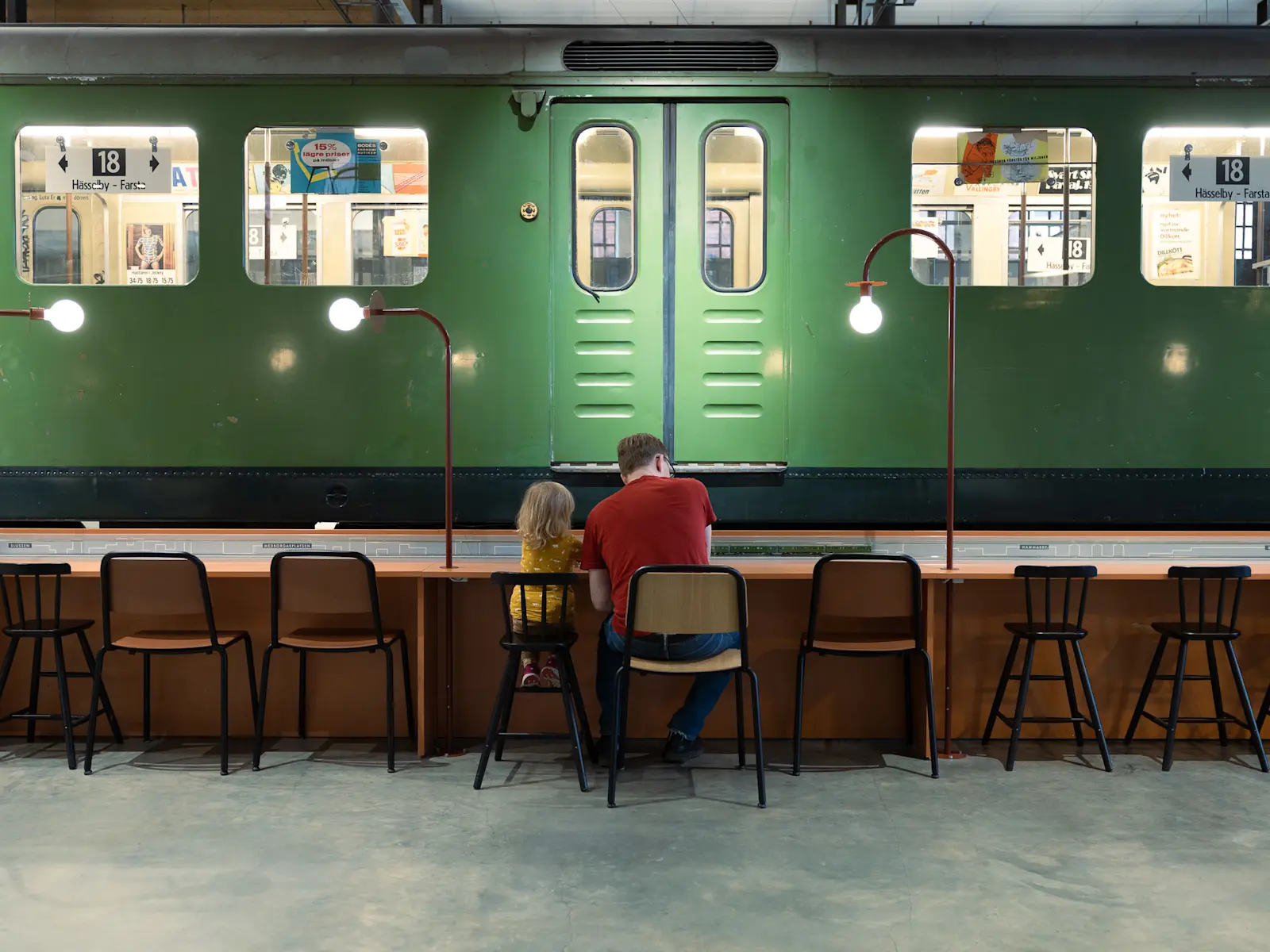 Ett långt cafébord framför en äldre grön tunnelbanevagn. Ett litet barn med axellångt blont hår och gulmönstrad tröja sitter bredvid en man i 30-årsåldern. Mannen bär en röd t-shirt och mörkblåa jeans. De sitter på varsin stol. I bild ser vi ett flertal trästolar, både svarta barnanpassade samt brunorangea med svarta ben.