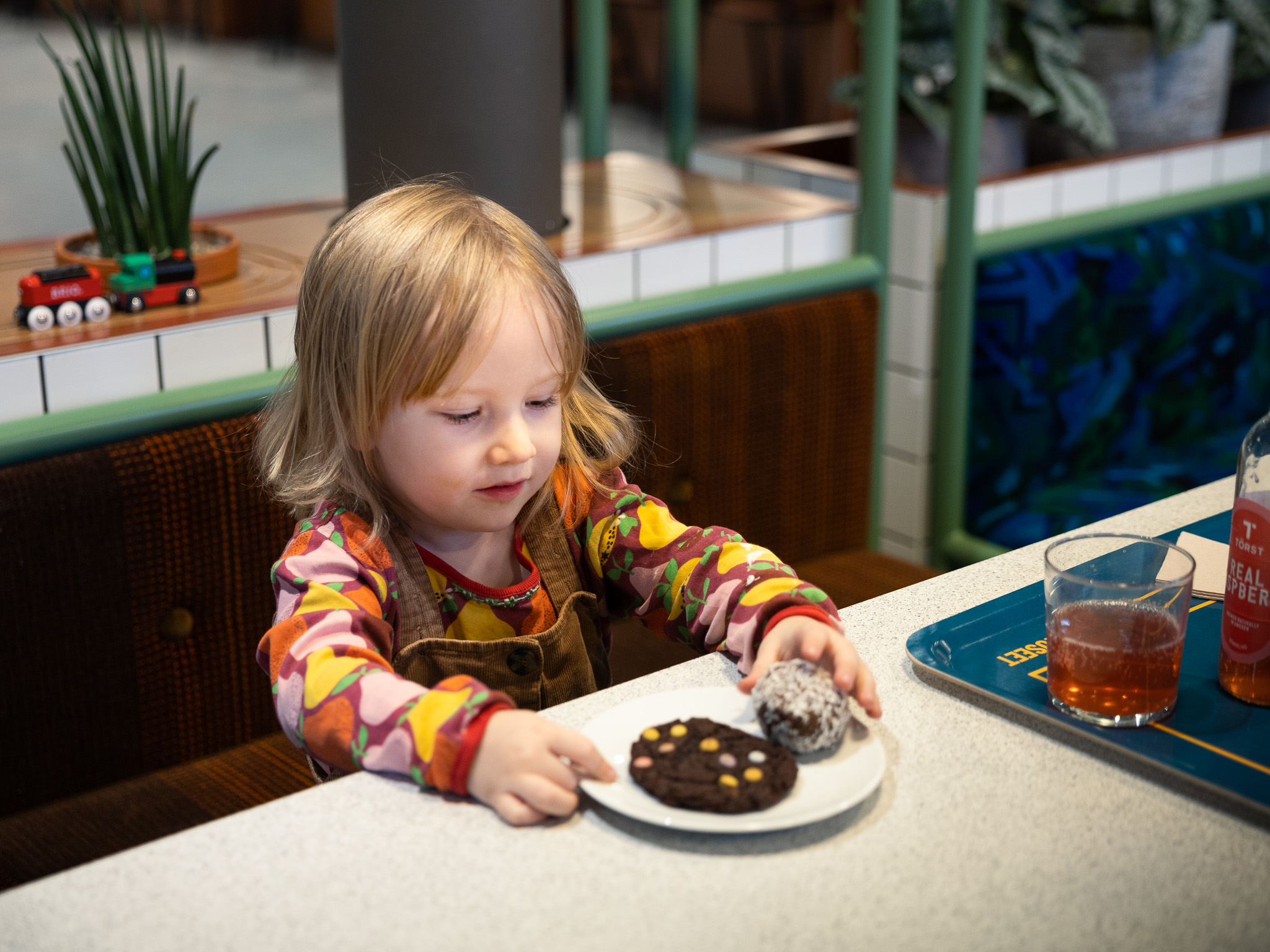 Ett litet barn i 3-årsåldern sitter i ett av caféets bås. Bänken barnet sitter på är ett återvunnet brunt tyg från ett äldre SL-fordon. Barnet har en brun tröja med fruktmönster på samt ett par bruna hängselbyxor. Barnet har en tallrik med en chokladcookie med chokladknappar i olika färger på samt en chokladboll. Till höger ser vi en blå bricka med gula detaljer på samt ett glas med rosafärgad läsk i. En glasflaska med samma innehåll står bredvid glaset. Bakom barnet ser vi en tågbana samt två stycken leksakståg.