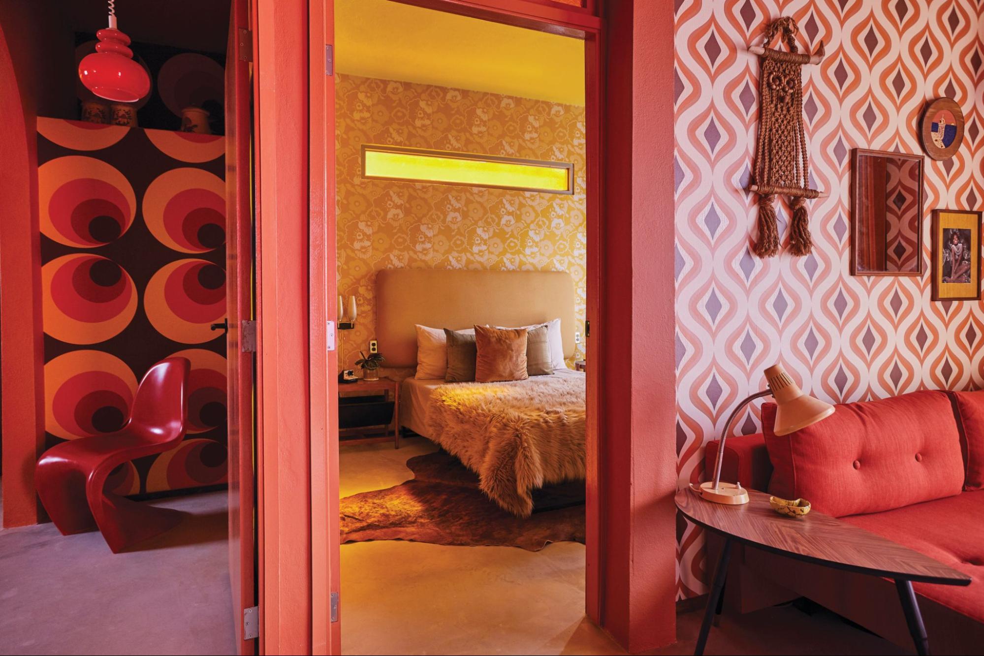 A vibrant retro-style suite featuring 1970s geometric wallpaper, a red curved Panton chair, a cozy bedroom visible through an open doorway, and a living area with a red sofa and vintage table lamp.