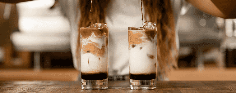A woman pours espresso into two tall glasses of milk to make iced coffees 