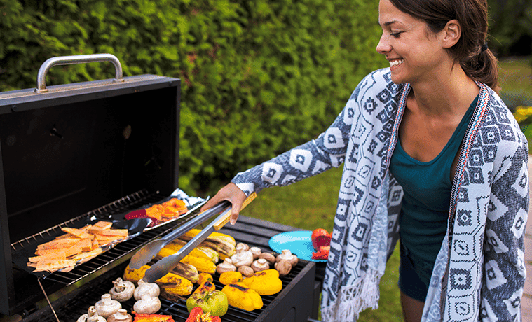 A young woman uses tongs to turn a colourful array of vegetables cooking on an outdoor barbecue
