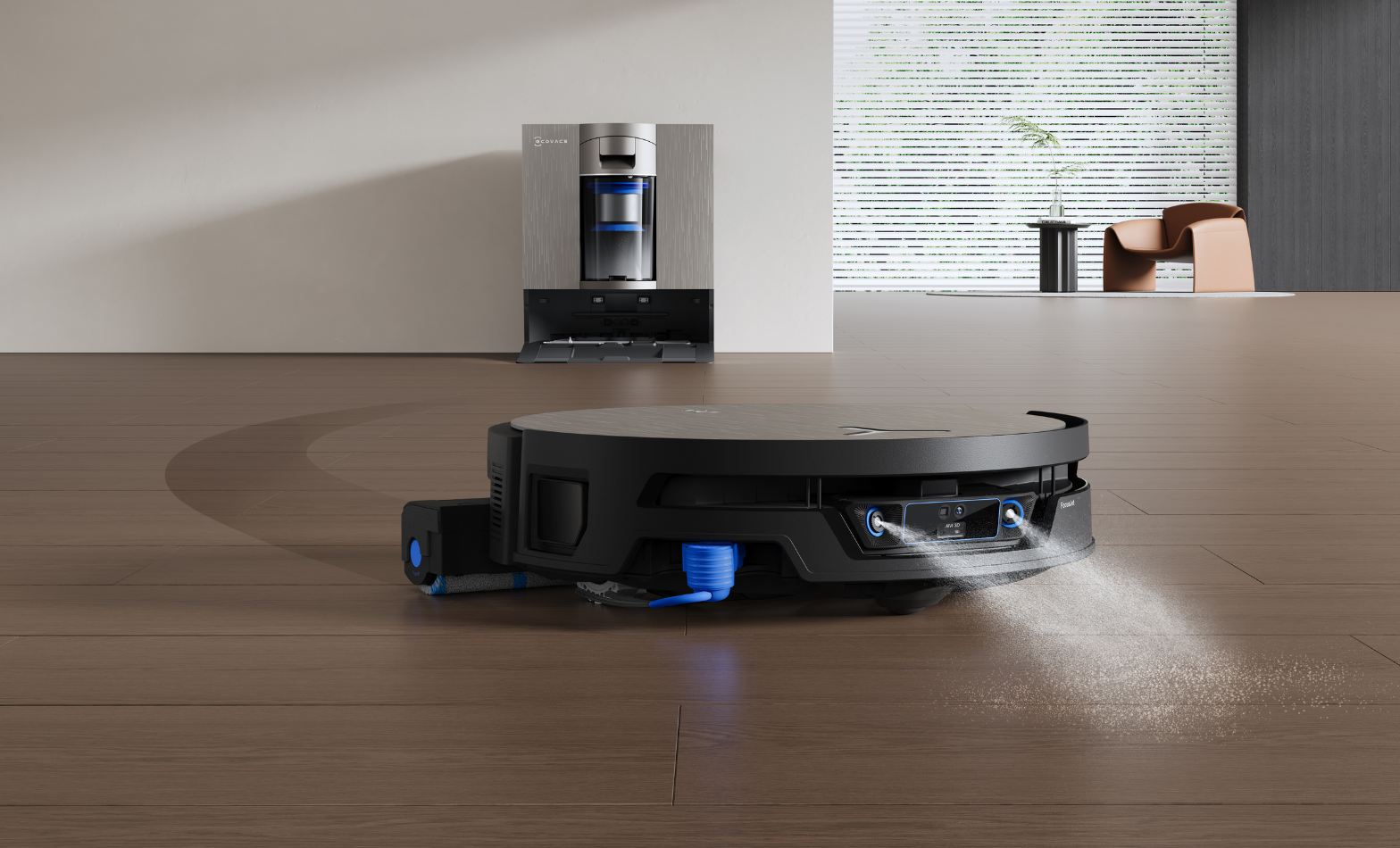 Close-up of a black ECOVACS robot vacuum cleaning a wooden floor, spraying a fine water mist from its underside with its extendable side mop arm reaching out to the left. Its tall silver docking station sits against the wall behind it in a bright, modern living room.