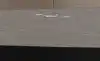 Close-up of a black ECOVACS robot vacuum cleaning a wooden floor, spraying a fine water mist from its underside with its extendable side mop arm reaching out to the left. Its tall silver docking station sits against the wall behind it in a bright, modern living room.