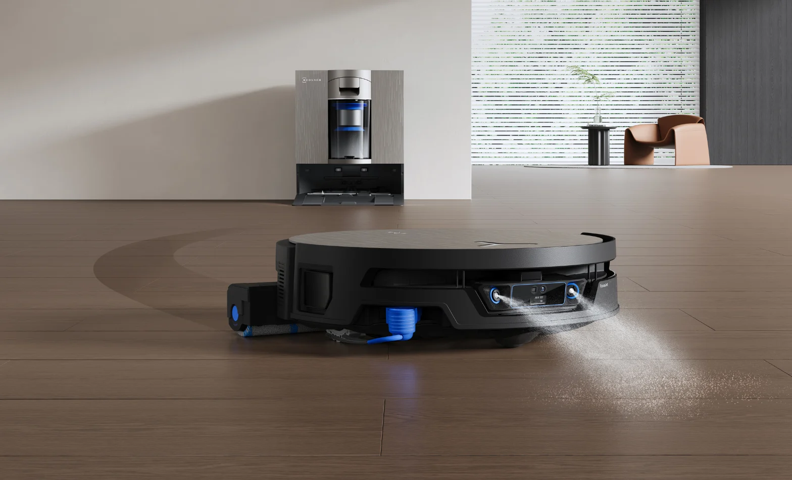Close-up of a black ECOVACS robot vacuum cleaning a wooden floor, spraying a fine water mist from its underside with its extendable side mop arm reaching out to the left. Its tall silver docking station sits against the wall behind it in a bright, modern living room.