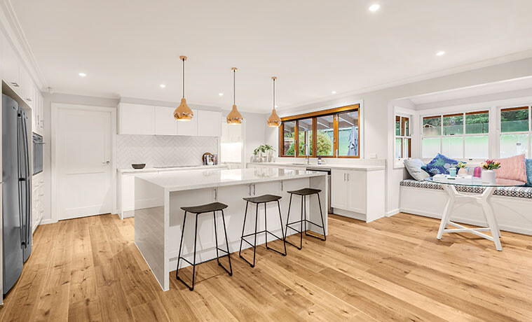Bright open plan kitchen with black stools timber floors and a trio of brasslook pendant lights above a white kitchen island