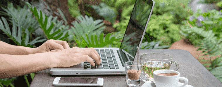 A person works on their laptop outside with a freshly brewed tea and coffee next to them