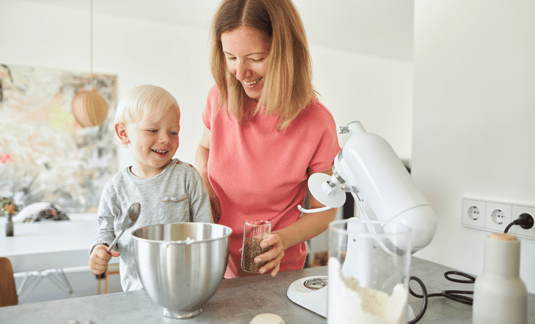A mother using a stand mixer while baking in the kitchen with her blondehaired toddler son