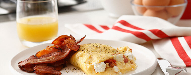 Bacon and omelette on a white plate with eggs orange juice and frypan and cooktop in the background