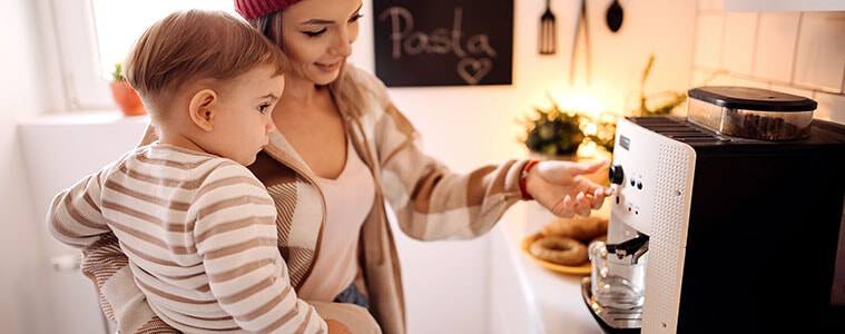 A mother holds her baby on her hip while turning on her coffee machine with her other hand
