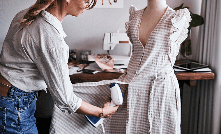 A woman uses a garment steamer to remove wrinkles from a dress on a mannequin in her sewing room at home