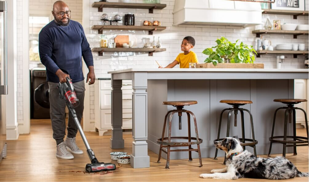 A father vacuums up his dogs spilled kibble while his young son watches