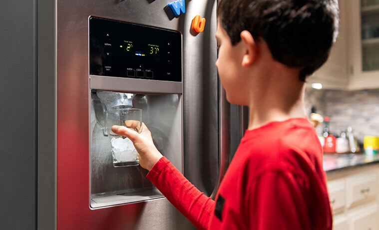 A young boy holds his cup to the iced water dispenser on the door of his fridge