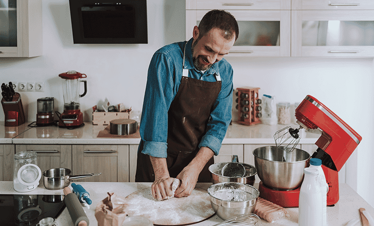 A man smiles and wears an apron while kneading a sweet pastry dough at his kitchen bench