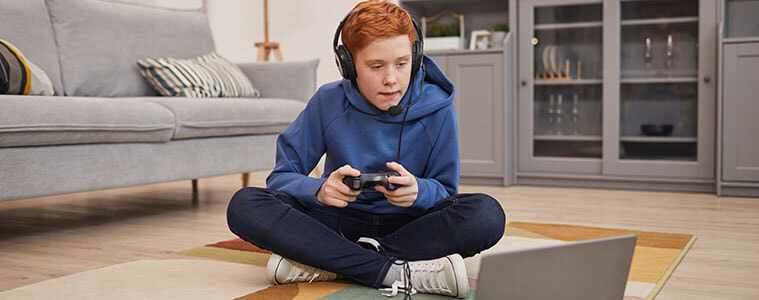 Boy sitting on the floor of his living room wearing a gaming headset and holding his controller which is plugged into a laptop