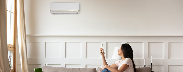 A woman sitting on the couch turns on her wallmounted air conditioner using a remote control