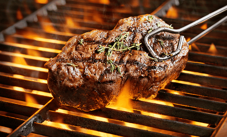 A closeup of a thick sirloin steak with fresh thyme above the flames of a barbecue grill