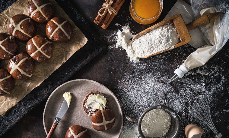 Overhead view of a dark stone kitchen bench dusted with flour with a buttered hot cross bun on a plate piping bag whisk 