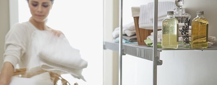 A woman takes a freshly folded white towel from the basket she is holding in her laundry with a neat and tidy shelving unit 
