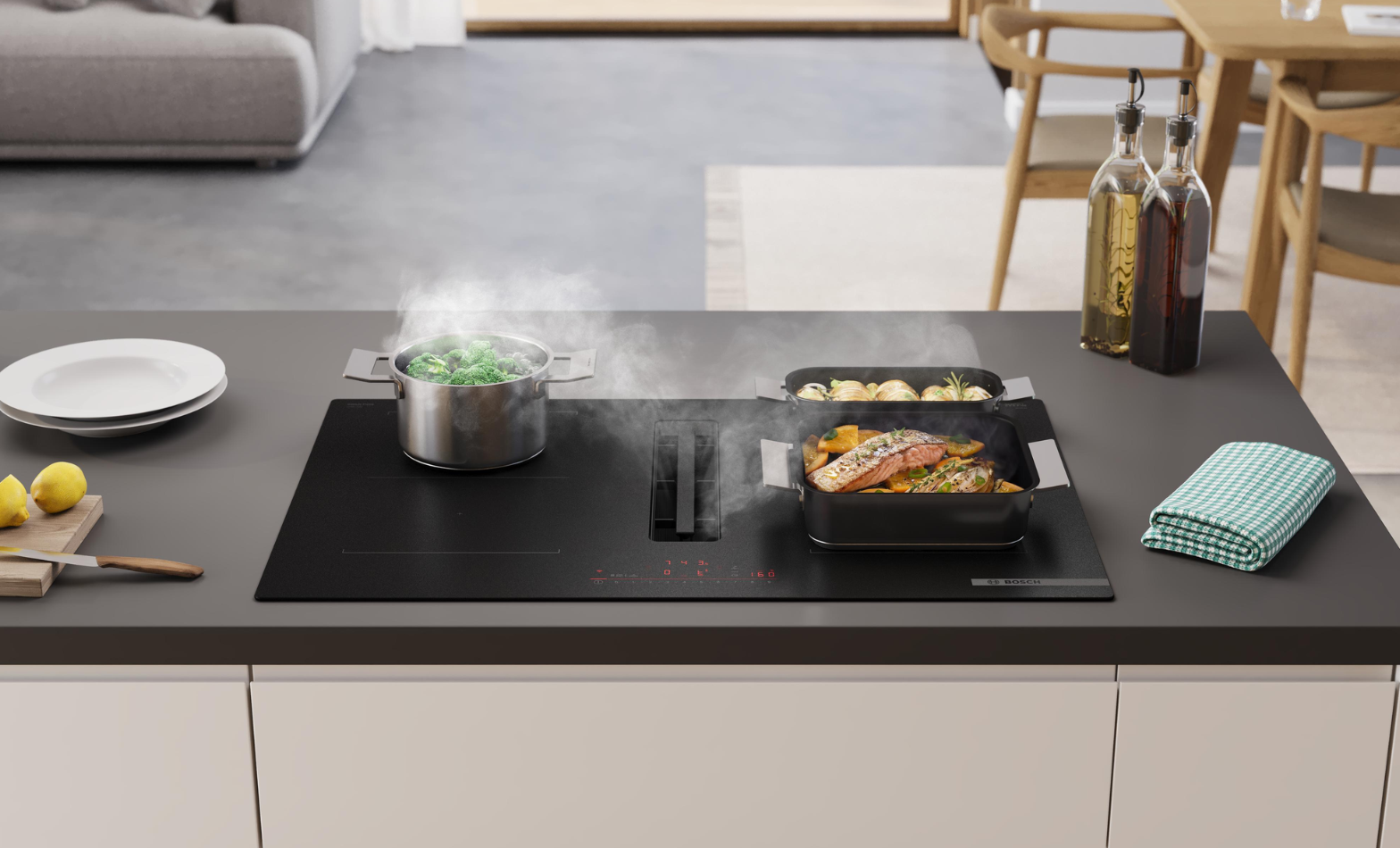 Bosch black induction cooktop with built-in downdraft ventilation, cooking salmon and steaming broccoli on a kitchen island.