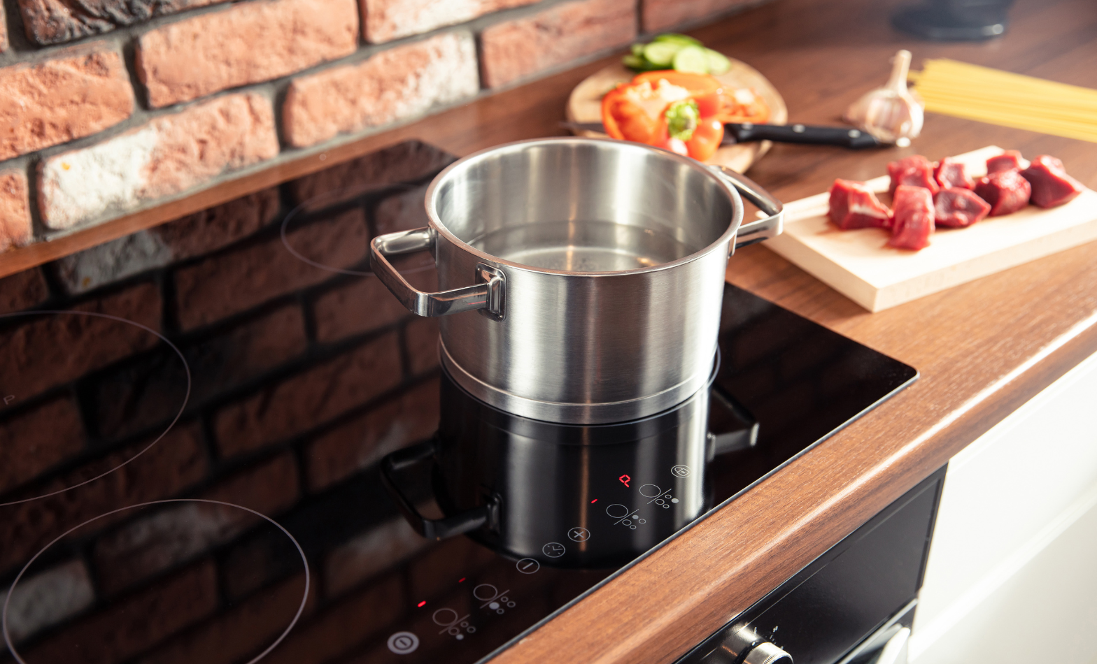 Stainless steel pot heating on a black induction cooktop with red digital touch controls, set in a kitchen with a timber benchtop, exposed brick wall, and fresh ingredients including beef, capsicum and garlic on a cutting board.