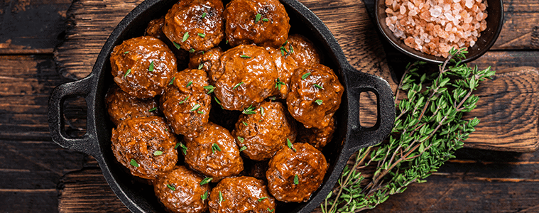Meatballs in a cast iron serving dish on a rustic timber table with a bouquet of herbs and a bowl of pink salt flakes 