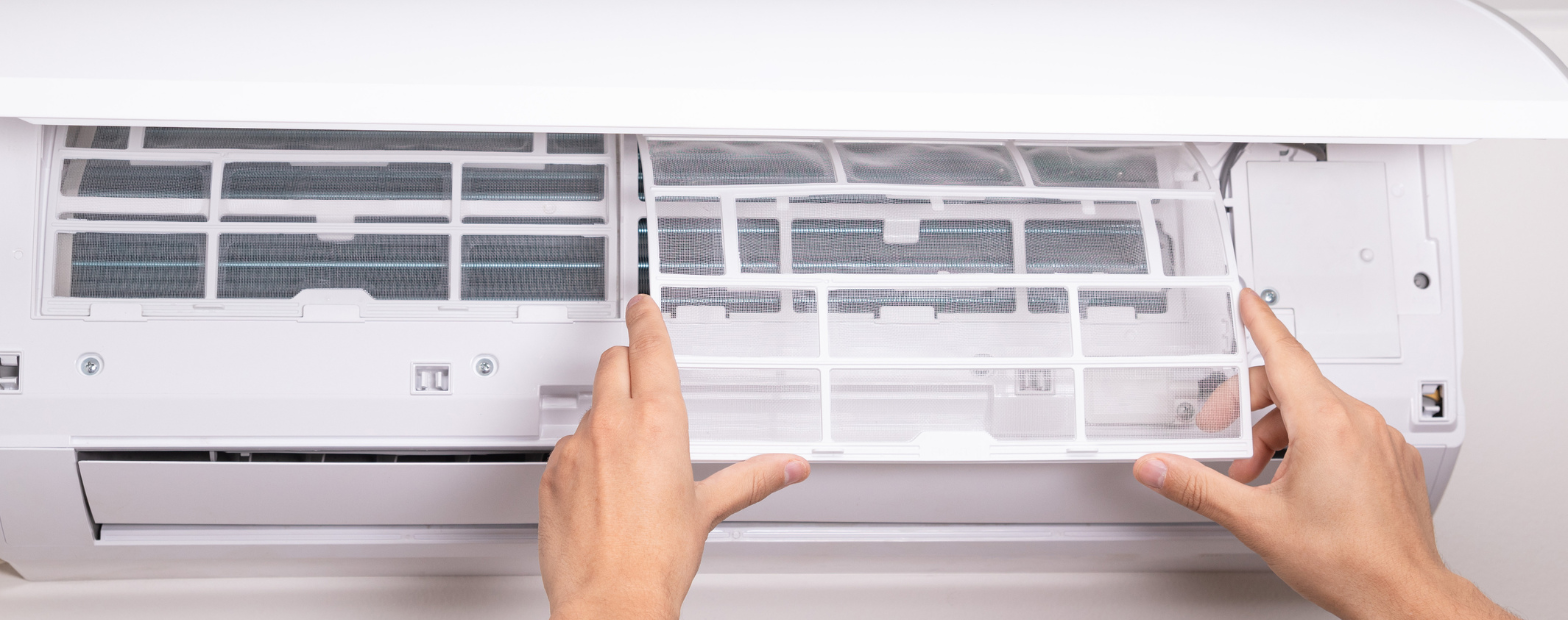 Hands sliding a mesh filter out of an open wall-mounted split system air conditioner.