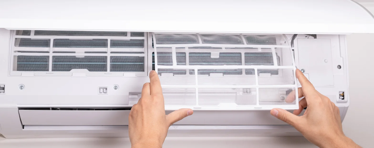 Hands sliding a mesh filter out of an open wall-mounted split system air conditioner.