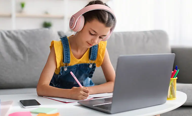 Primary schoolaged girl writes in her text book on the living room coffee table with her laptop open in front of her