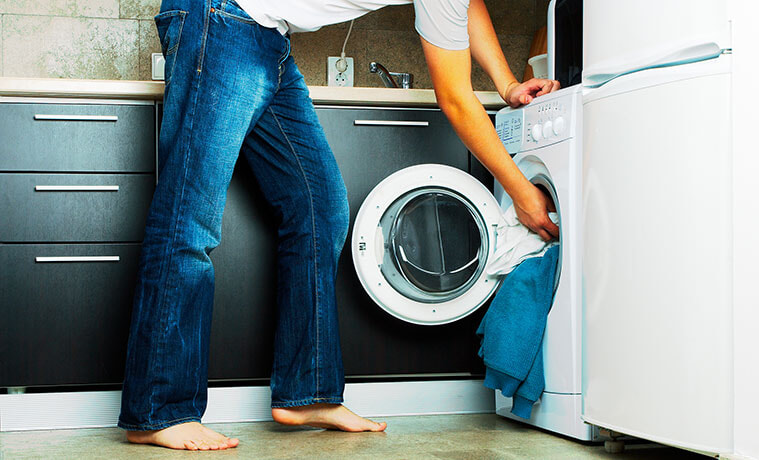 Waistdown view of a man in blue jeans loading white bed linen into a front loader washing machine