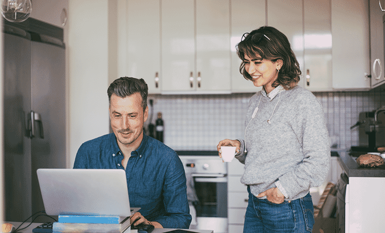 Smiling man and woman in the kitchen at home with man at the table working on a laptop