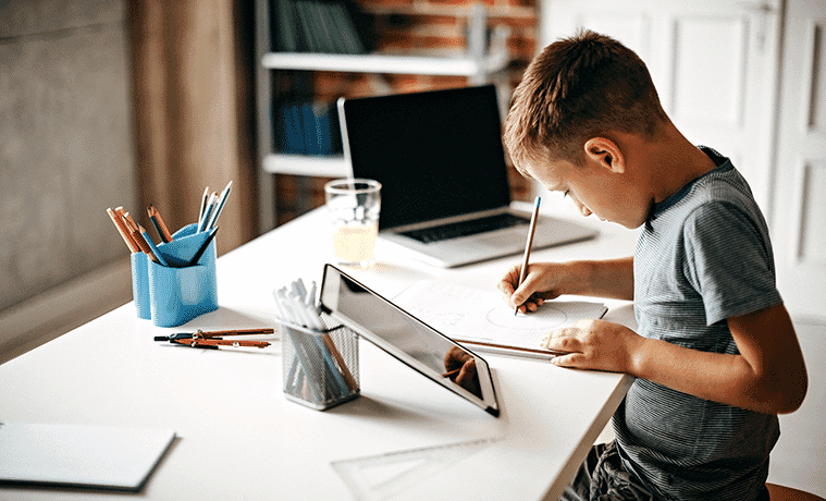 A primary school aged child does his homework while sitting at a desk with a tablet and laptop computer