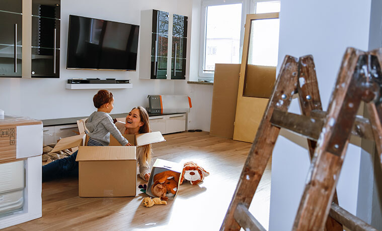 A mother and her young son sit on the floor of the living room surrounded by moving boxes and with a big screen TV on the wall