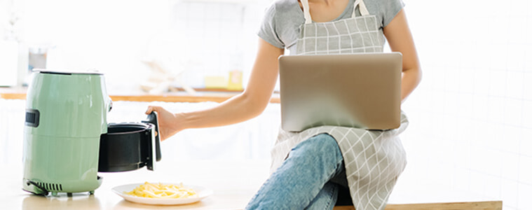 Woman sits on her kitchen bench with a laptop in her lap while removing snacks from her air fryer with her free hand