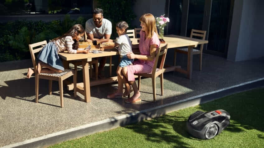 A young family have afternoon tea and play a card game while a Husqvarna robot lawn mower mows their lawn
