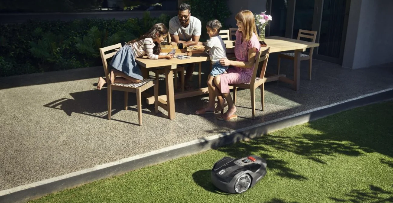 A young family have afternoon tea and play a card game while a Husqvarna robot lawn mower mows their lawn