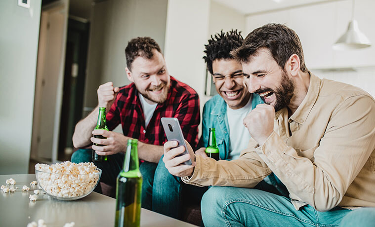 Three male friends sit on the couch drink beer and cheer as they watch a match on the screen of a smartphone