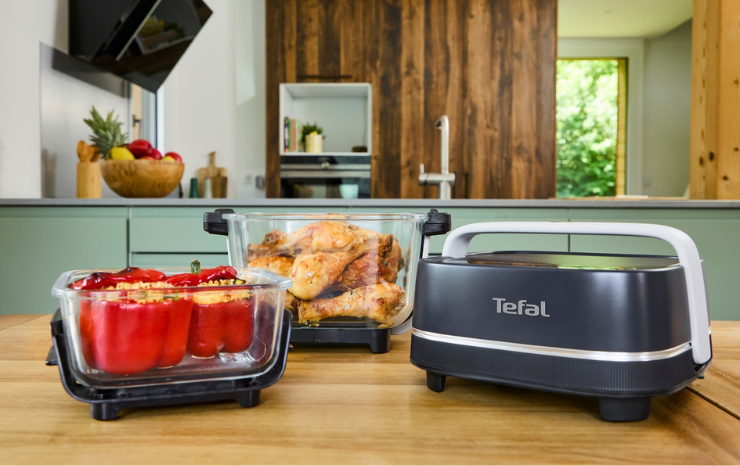Tefal Crisp & Go glass air fryer with its heating lid beside two glass bowls, one with roast chicken drumsticks and one with stuffed red capsicums, on a wooden kitchen bench.