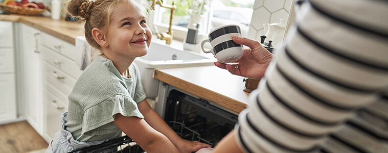 A daughter smiles at her mum while helping her to load cups and plates into the dishwasher