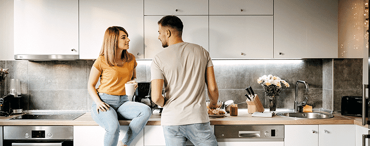 Woman sits on the kitchen bench to talk to her partner while he makes lunch in their modern white galley kitchen with an under bench oven and sleek electric cooktop 